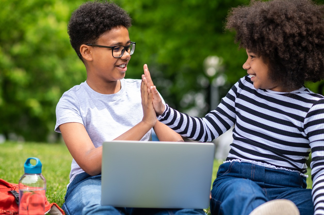 Two children sitting on grass with a laptop preparing for an online programming camp