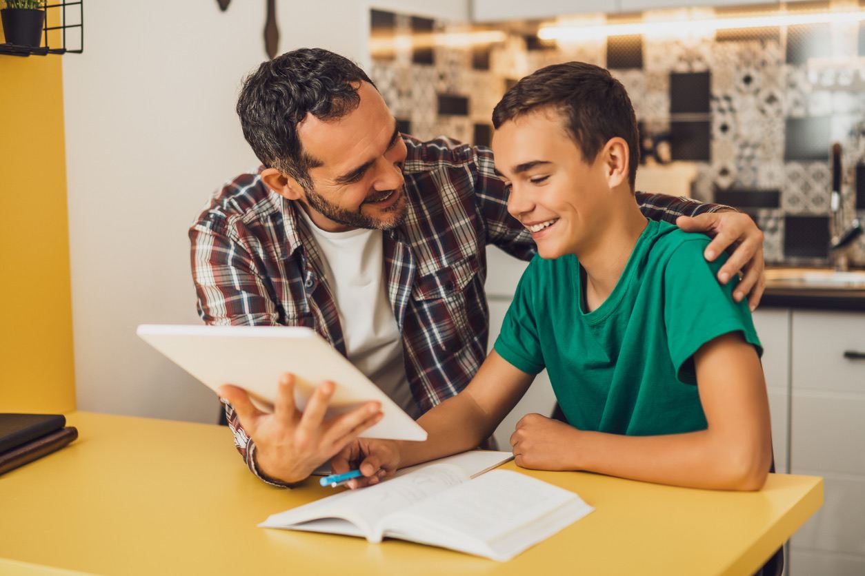 Father and son sitting at a table, looking at a tablet and smiling.