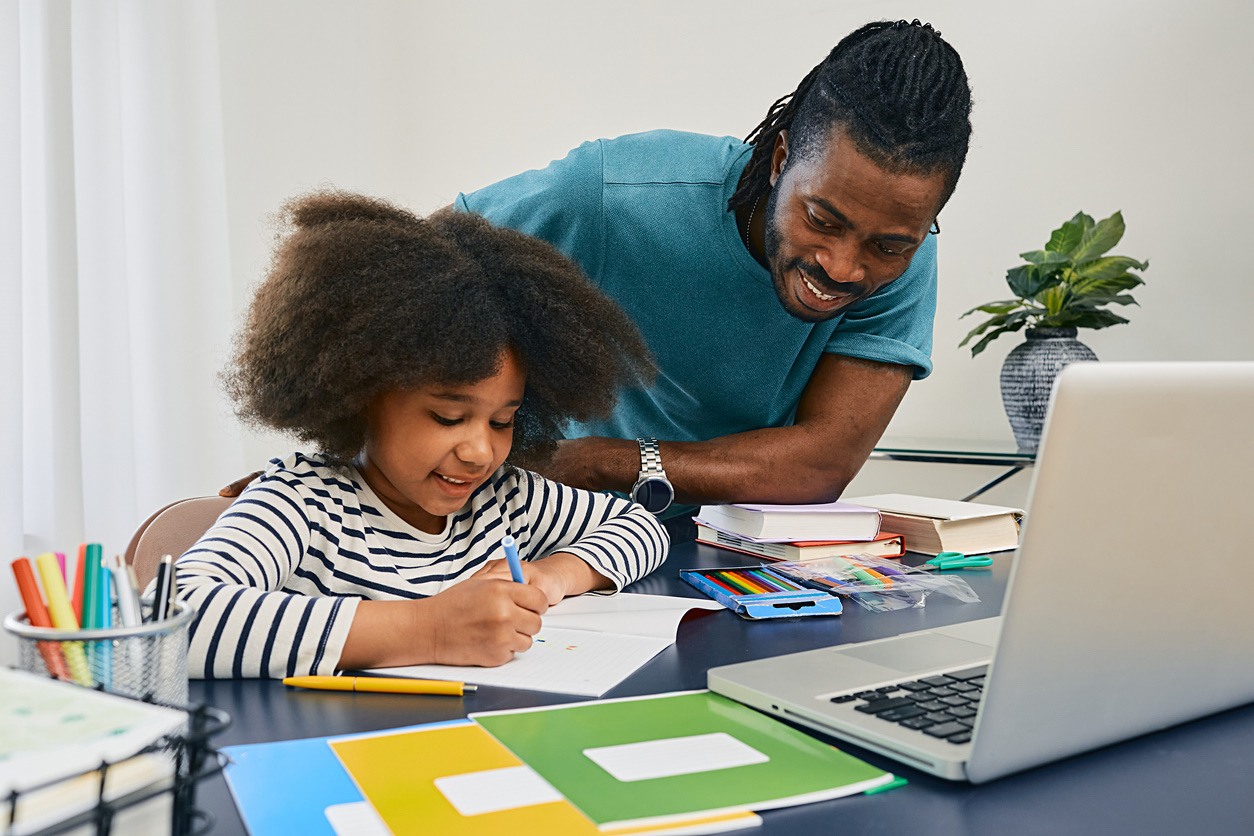 Father supporting his daughter during a writing activity at a desk with a laptop.