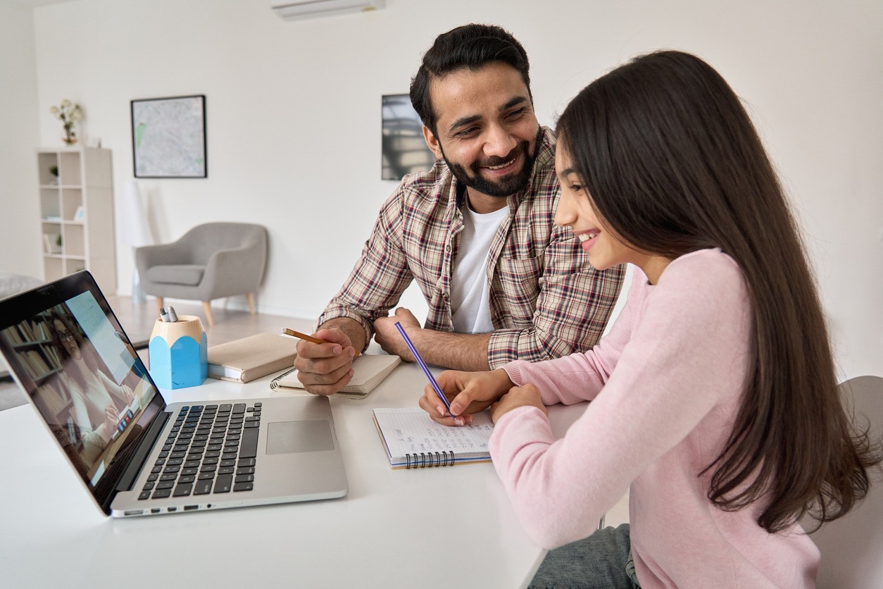 Father and daughter smiling while studying together using a laptop at home.