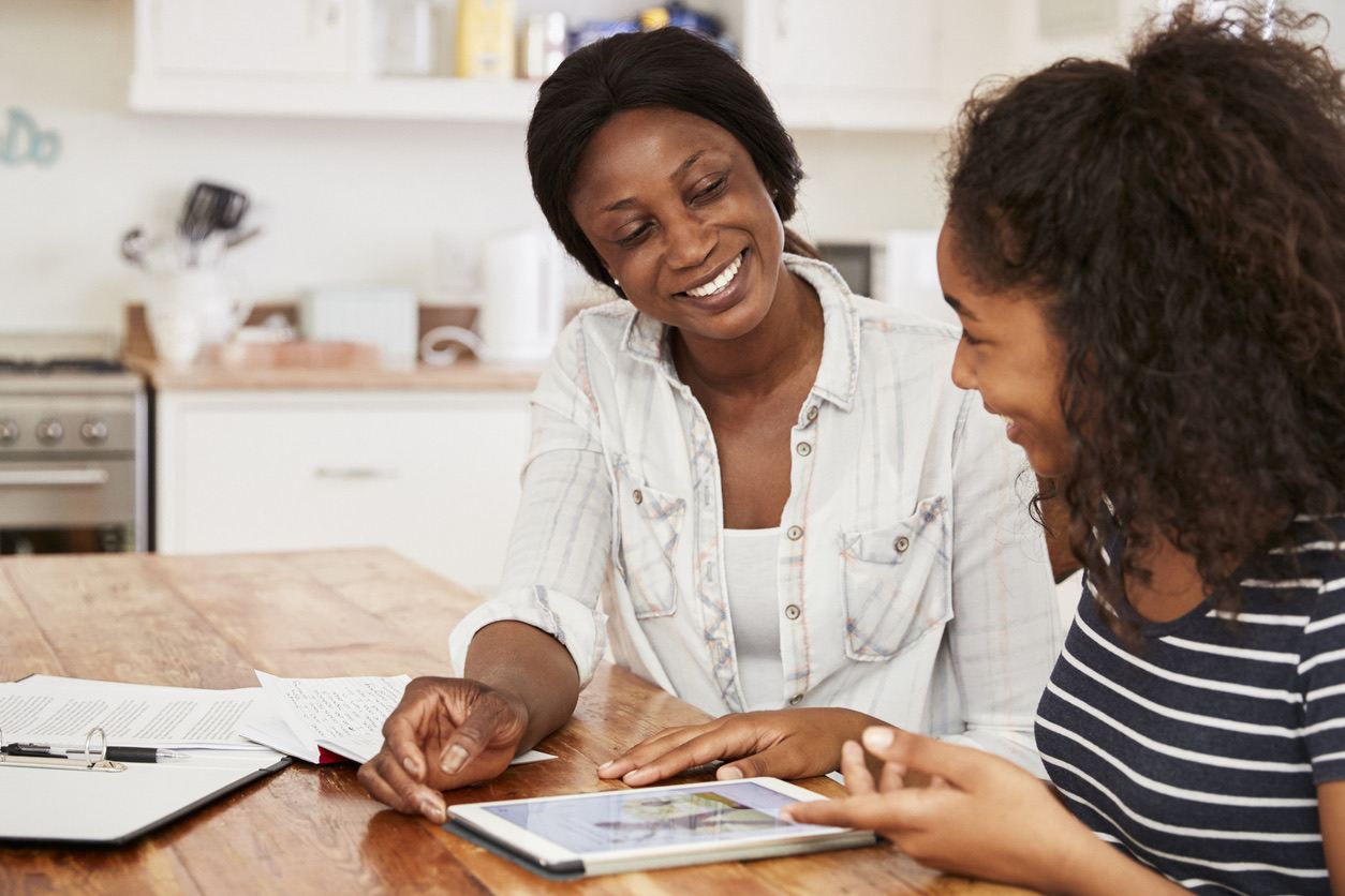 Mother and daughter smiling while learning on a tablet at the kitchen table.