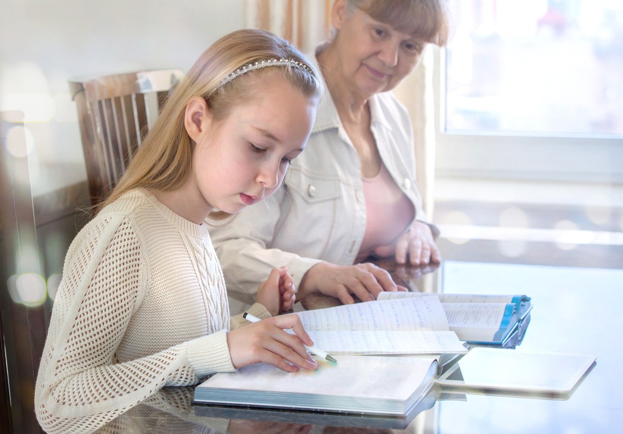 Young girl learning to write with an elderly woman at a table.